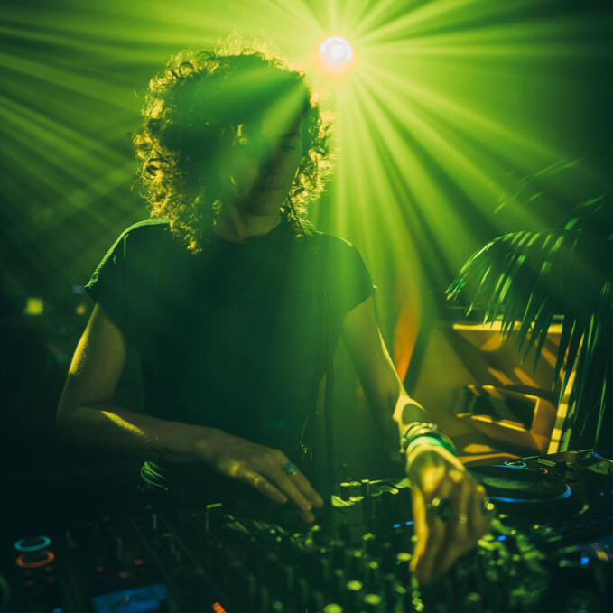 DJ Lamírla performs behind the decks under bright green beams of light, focused on the mixer during her set.