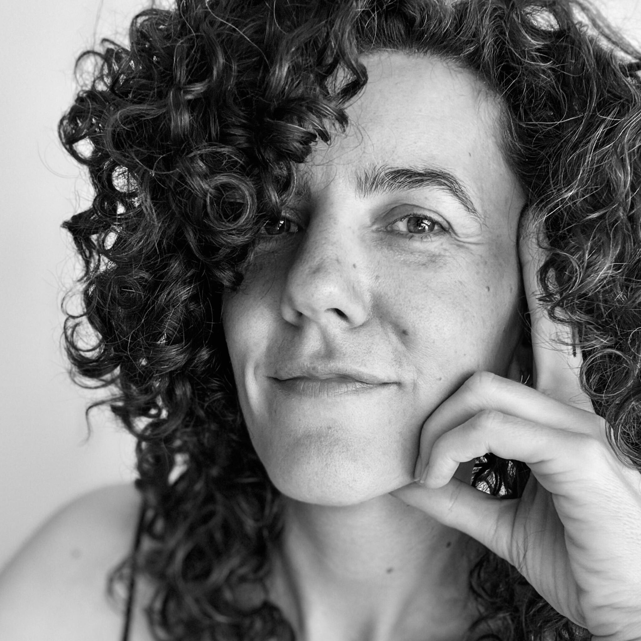 Black-and-white portrait of DJ Lamírla looking into the camera with a subtle smile, resting her chin on her hand. Her curly dark hair partly covers one side of her face.