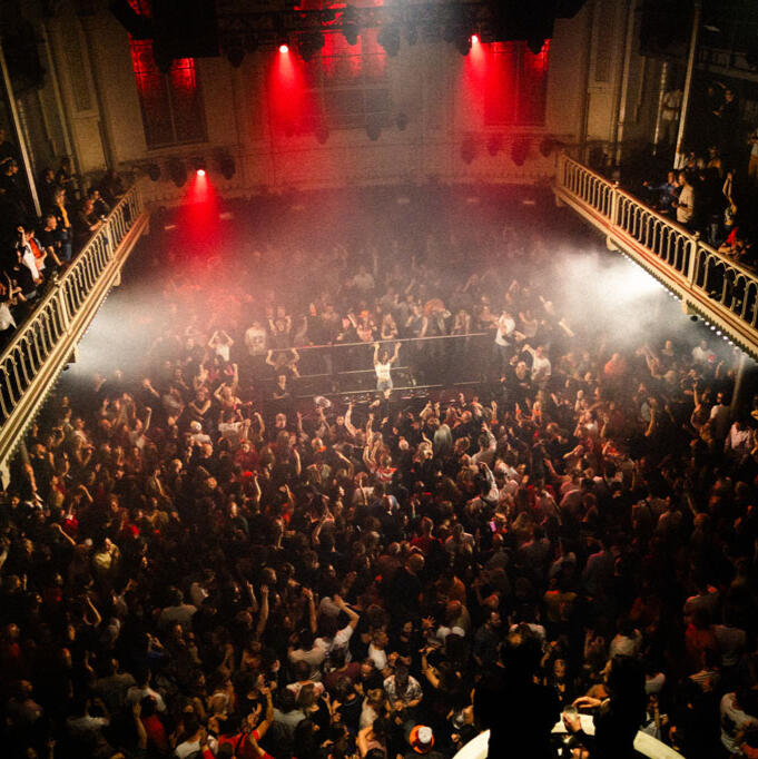 Wide view of a sold-out Paradiso Amsterdam with a crowded dance floor. DJ Lamírla stands at the center behind the decks, surrounded by an energetic audience.