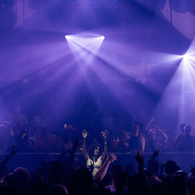 Lamírla DJ Performance Paradiso DJ Lamírla stands behind the decks in Paradiso Amsterdam with both hands in the air as a packed dance floor celebrates in front of her. Purple beams of light and haze fill the venue.