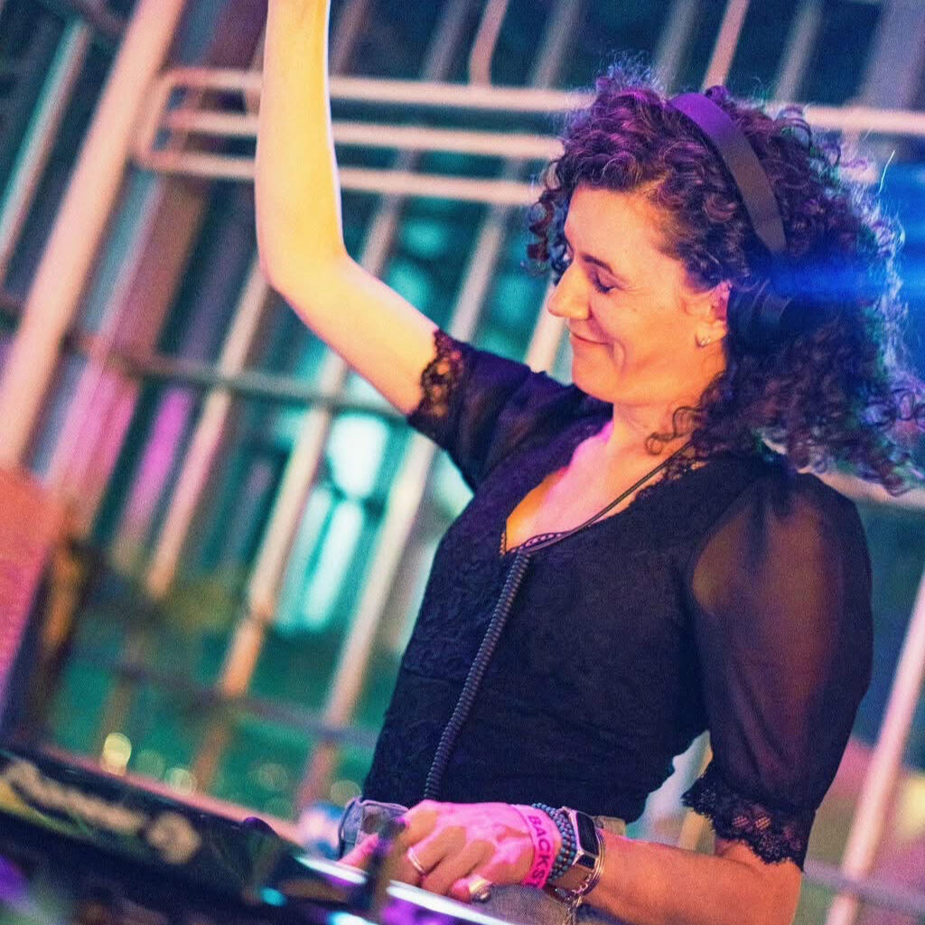 DJ Lamírla performs behind the decks with one arm raised and a smile on her face. Colorful lighting creates a vibrant festival atmosphere.
